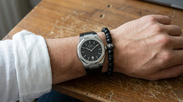 A man's wrist on a wooden desk wearing the Brother & Sisters Black Signature octagonal watch on black crocodile strap stacked with a Black Lion bead bracelet, men's accessory wrist-stacking trend