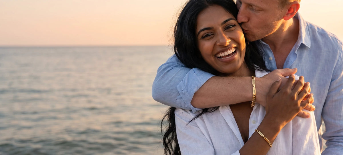A couple laughing on the beach at golden hour, each wearing a matching gold bracelet